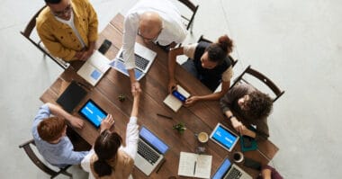 Overhead view of a diverse group of six professionals sitting around a rectangular table, engaging in discussion on business strategies and working on laptops.