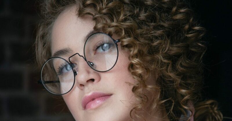 Close-up portrait of a person with curly hair and round glasses, looking upward with a neutral expression, set against a dark, blurred background. The introspective gaze hints at the journey of mastering FeetFinder or perhaps contemplating the nuances of buying feet pics in today's digital age.