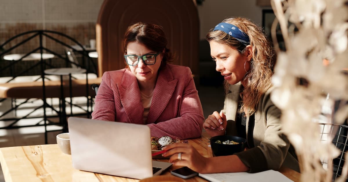 Two women collaborating on a Shopify project using a laptop in a sunlit cafe.