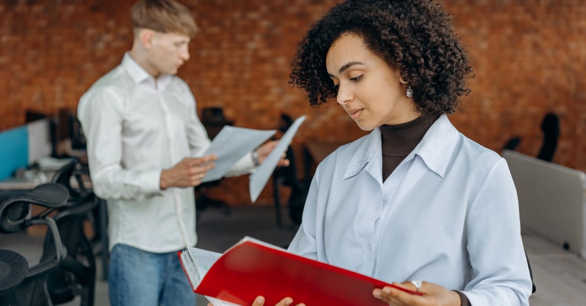 A woman in a white coat reviews demand forecasting documents in a folder in a modern office setting, with a man in the background reading papers.