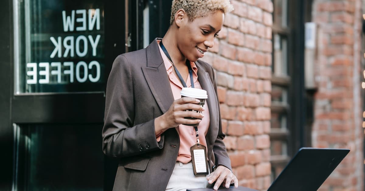 A professional woman smiling, holding a coffee cup and using a laptop outside a cafe with an "Ecommerce Tools" sign in the background.