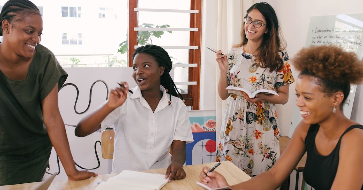 Four women engaging in a lively discussion around a table with books and papers, focusing on business essentials, in a brightly lit classroom.
