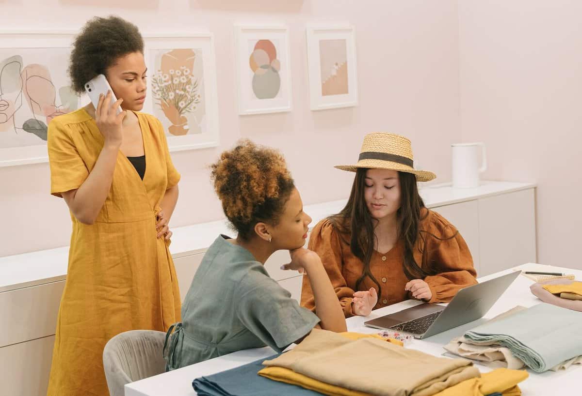 Three women in a boutique, one on the phone and two discussing digital marketing strategies over a laptop, surrounded by clothing and art.