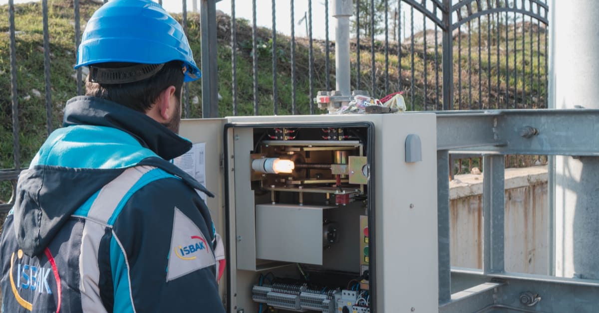 A professional technician in a blue helmet and vest is inspecting the interior of an open electrical cabinet outdoors.