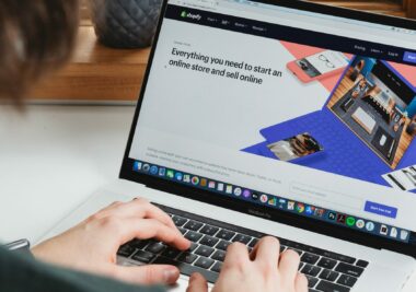 A person types on a laptop displaying the Shopify website homepage, which promotes starting an ecommerce business and selling products online.