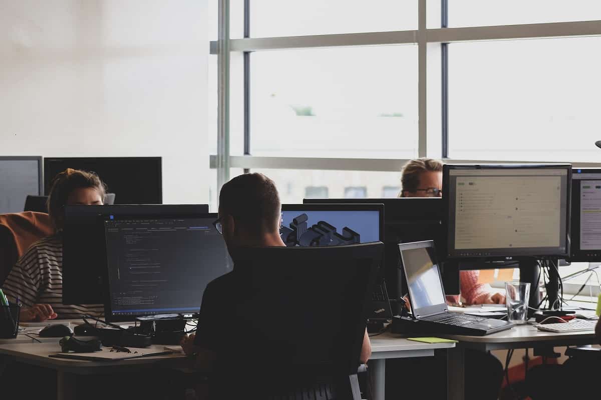 Three office workers at desks with computer monitors, one displaying online promotions, in a brightly lit room with large windows.
