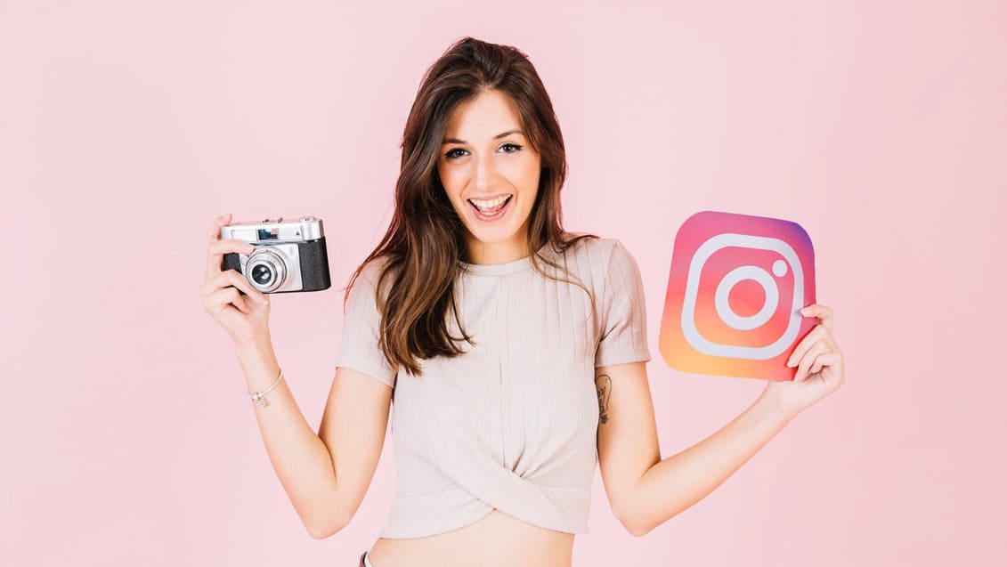 A smiling person holds a camera in one hand and an Instagram logo cutout in the other, standing against a pink background, showing they have mastered the Instagram game.