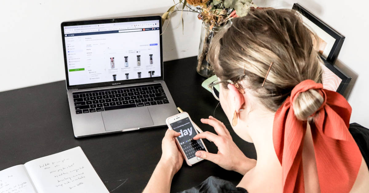 Woman with a red hair tie using a smartphone and laptop simultaneously at a black desk with various items, including a dried flower bouquet, focused on ecommerce best practices.