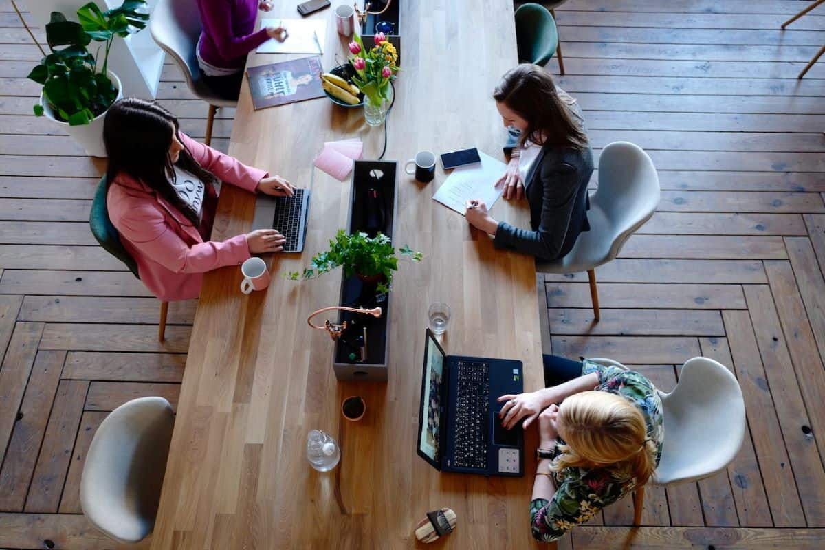 Three people working at a wooden table with laptops and notebooks; potted plants and assorted items are placed on the table. The setting appears to be an open, collaborative workspace focused on brand consistency.