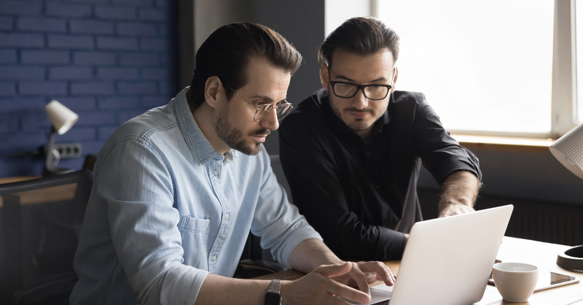 Two men wearing glasses and casual clothing are seated at a table, looking at a laptop screen together. One points at the screen while the other types on the keyboard, seemingly working on a SaaS platform for shoppable video.