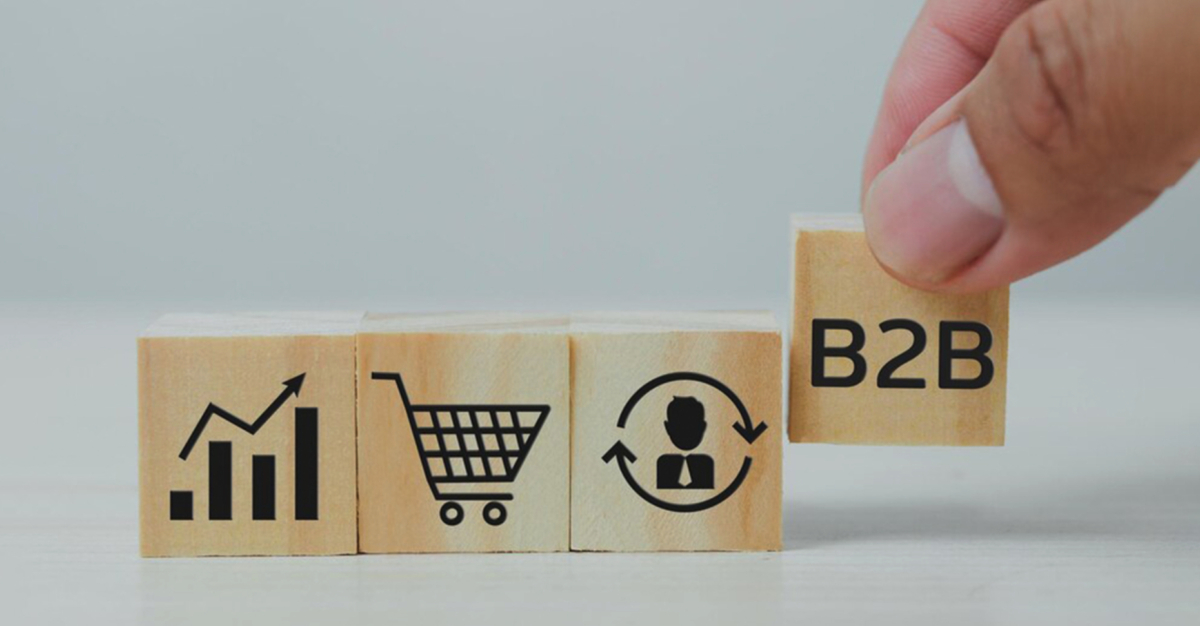 A hand arranges wooden blocks depicting business-related icons: a growth chart, a shopping cart, a customer relationship symbol, and a block labeled "B2B E-commerce.