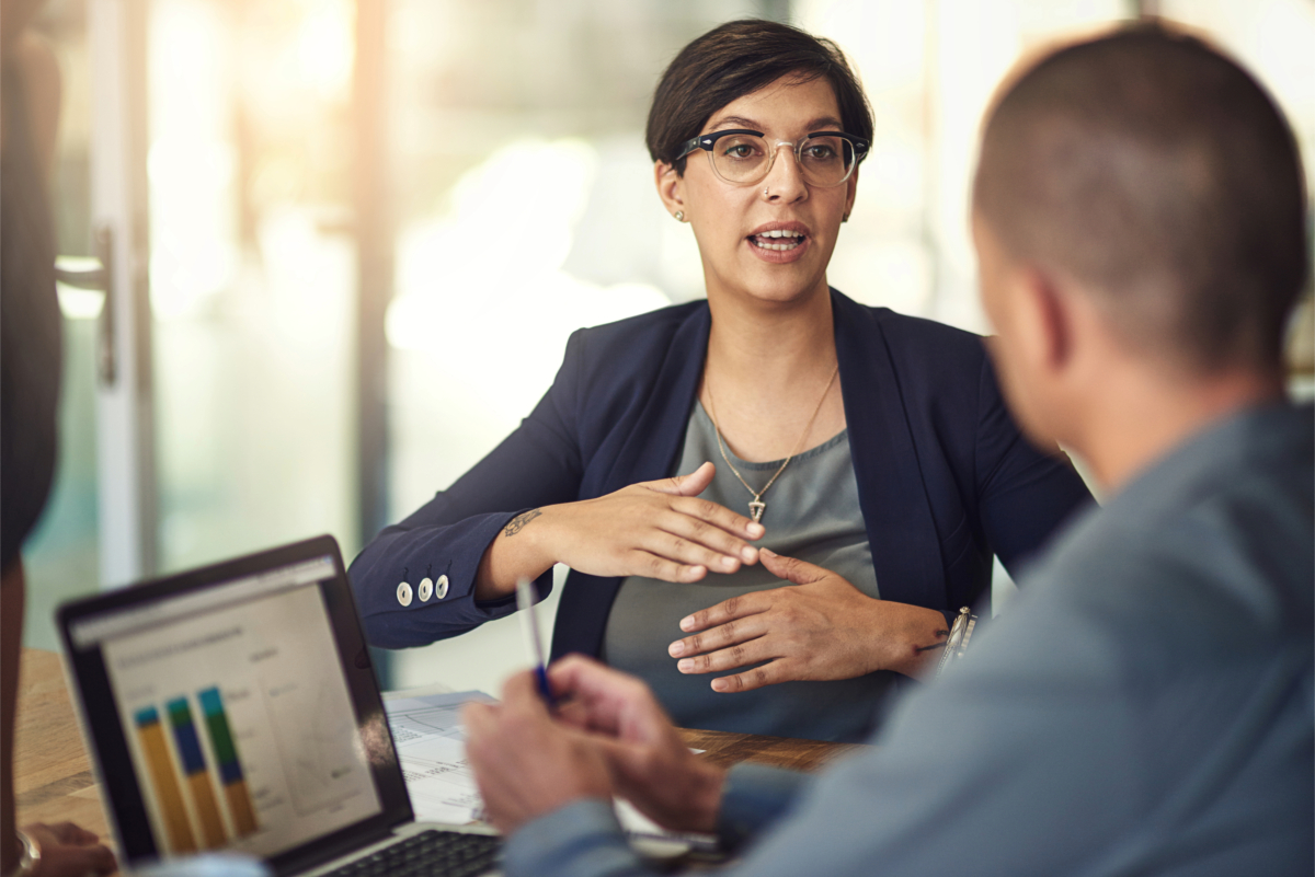 A woman wearing glasses and a blue blazer speaks and gestures during a business meeting, focusing on B2B sales strategies to boost results. Two colleagues are blurred in the foreground. A laptop displaying a bar chart is on the table.