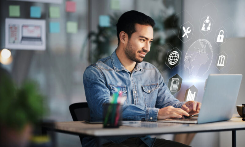 A man in a denim shirt works on a laptop in an office setting, with various digital icons floating around the screen, highlighting business tools and indicating tech-related activities.
