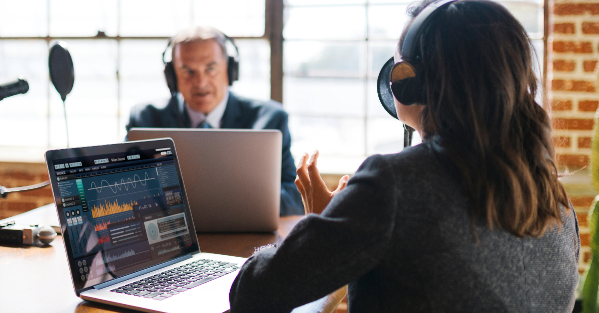Two people wearing headphones sit at a table with laptops, recording an important video podcast in a brick-walled room with large windows. Audio waveforms are visible on one laptop screen.