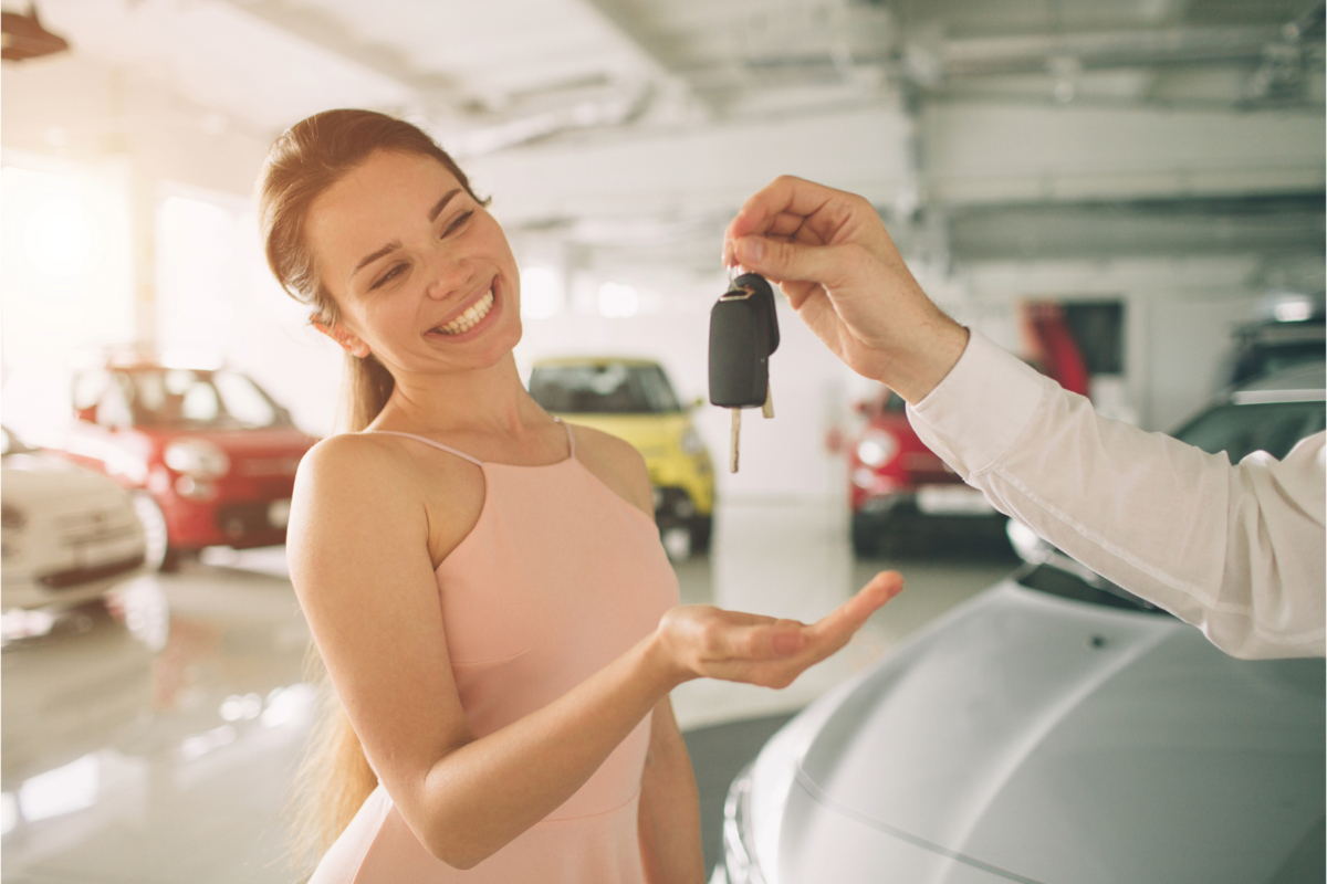 A person smiling as another hands over car keys in an indoor used car showroom, a moment that marks the successful culmination of a comprehensive buying guide.