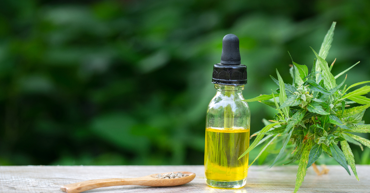 A dropper bottle with a yellow liquid is placed on a wooden surface next to a wooden spoon containing seeds and a hemp plant, capturing all you need to know about natural wellness.