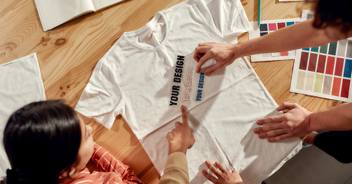 Two people customize a white T-shirt with different text designs on a wooden table, featuring design samples and color swatches—a perfect example of crafting commerce.