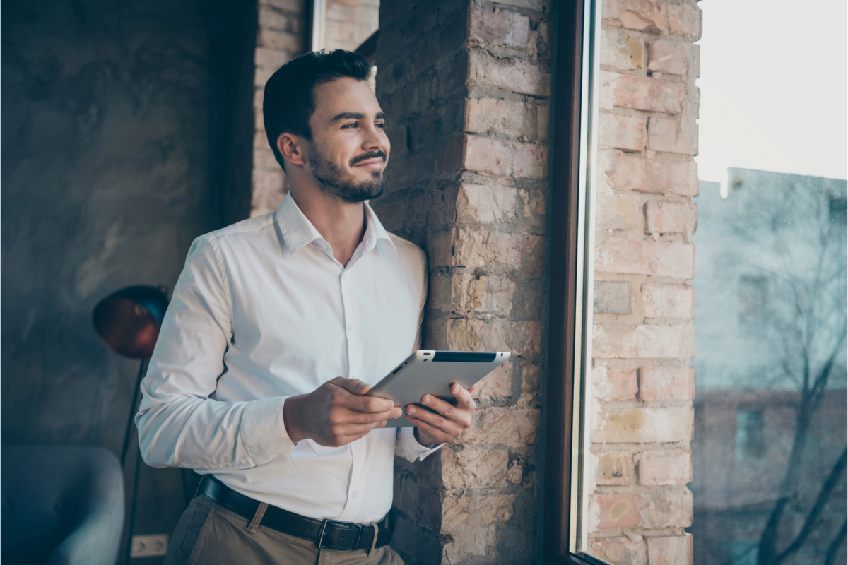 A man in a white shirt holds a tablet and looks out a window, smiling. He is leaning against an exposed brick wall inside a building, contemplating the financial freedom that comes with buying an ecommerce franchise.