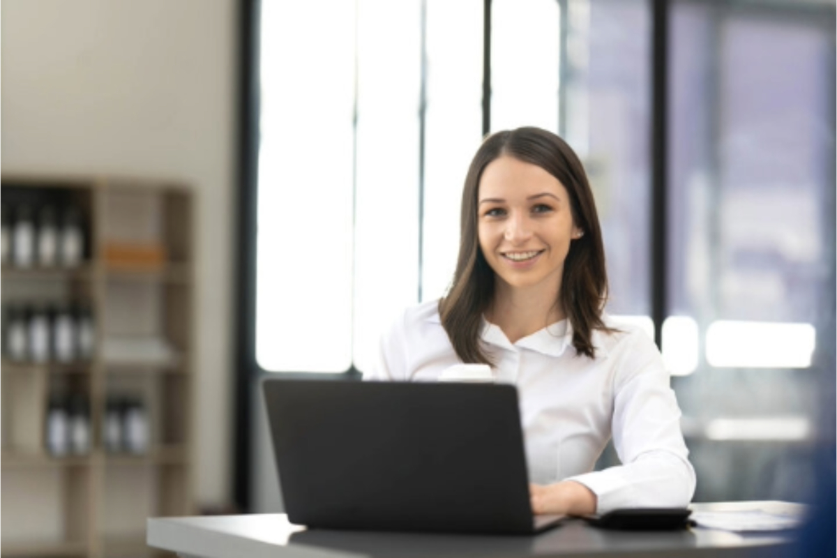 A woman sits at a desk with a laptop in front of her, smiling at the camera in a well-lit office environment, possibly working on the ultimate guide to creating a successful loyalty program for Shopify users.