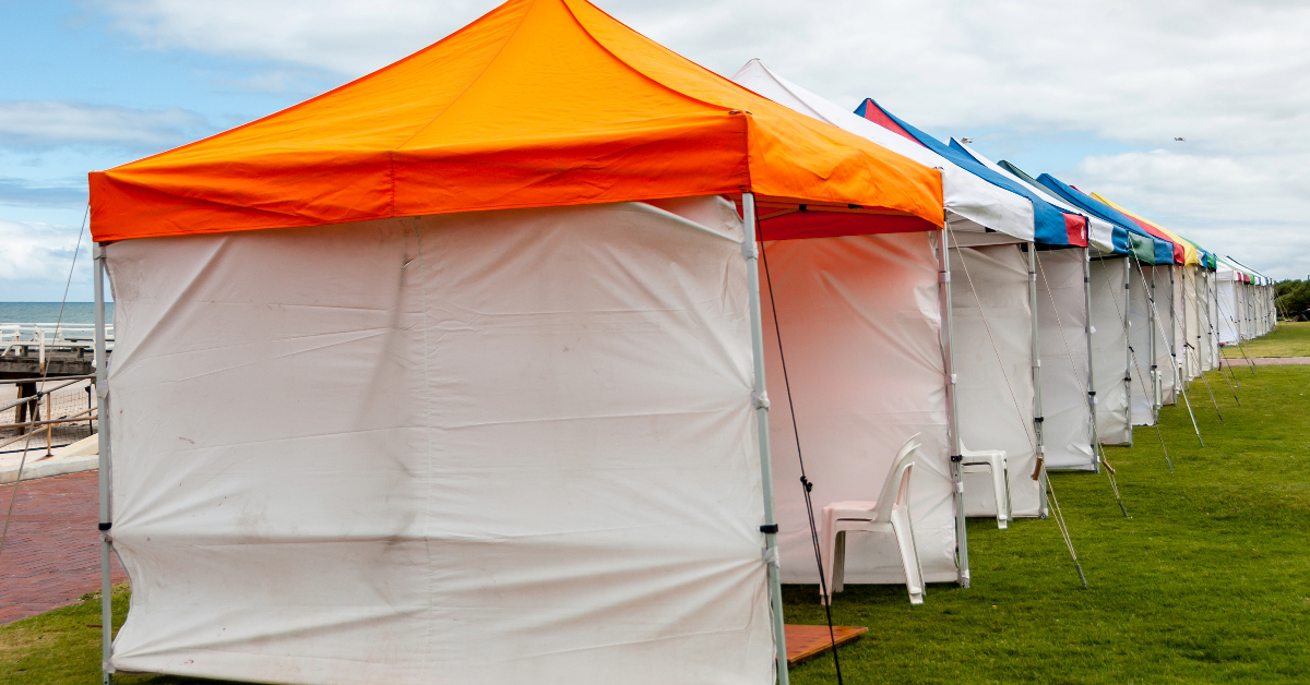 Several branded marquees with different colored tops are set up in a row on grass near a paved surface, with an empty white plastic chair in front of the nearest tent, creating an ambiance of outdoor luxury.