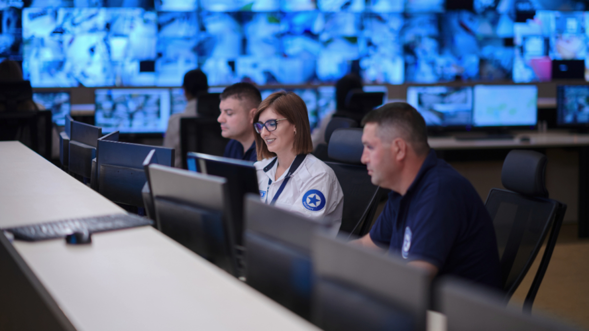 Three people in uniform monitor multiple screens in a control room. A woman in the center wears glasses, and two men are seen beside her, focusing on their screens. The team leverages ad ops software to optimize workflow and streamline ad operations efficiently.