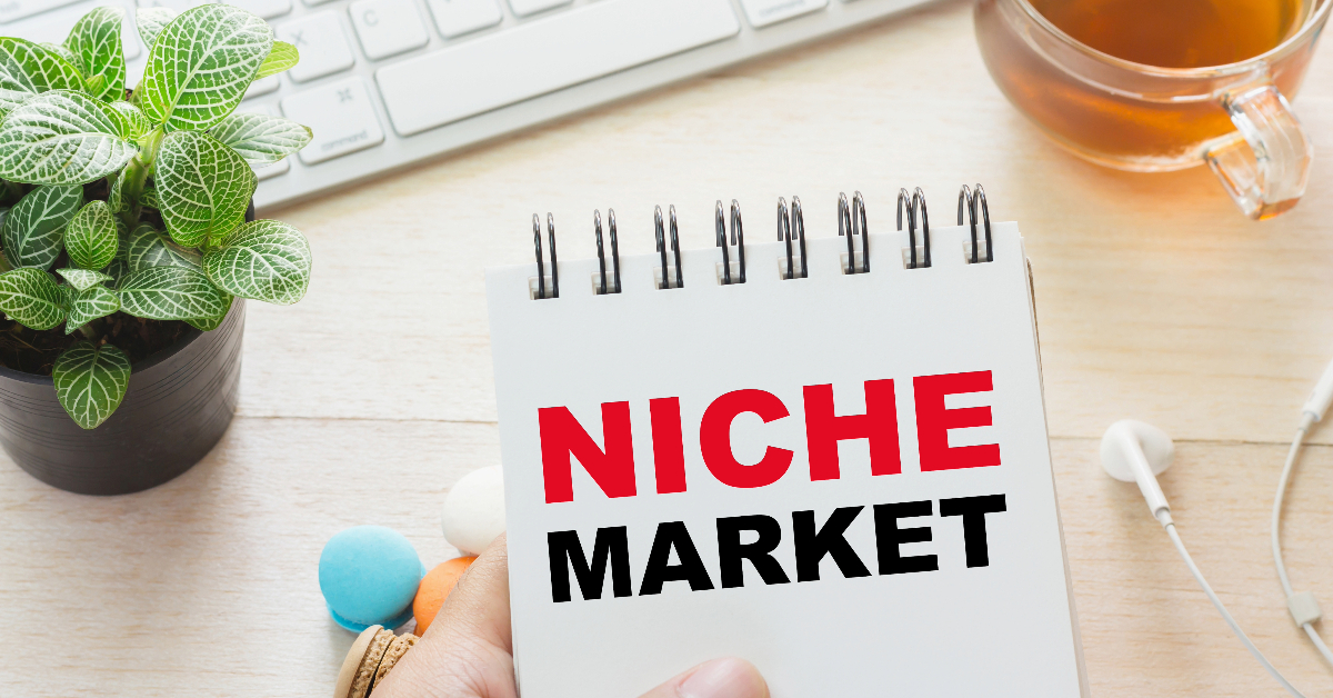 A notebook with the words "NICHE MARKET" in bold red and black letters, held in front of a desk with a plant, computer keyboard, tea, and earphones—perfect for your eCommerce or niche marketing strategies.