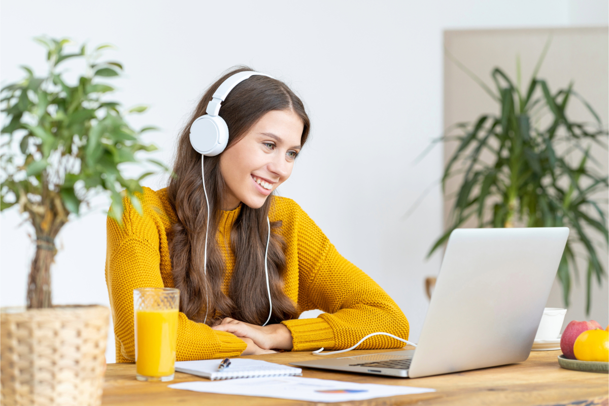 A woman in a yellow sweater wearing headphones is smiling at a laptop, perhaps researching side hustles. A notebook, pen, and glass of orange juice are on the table with plants in the background.