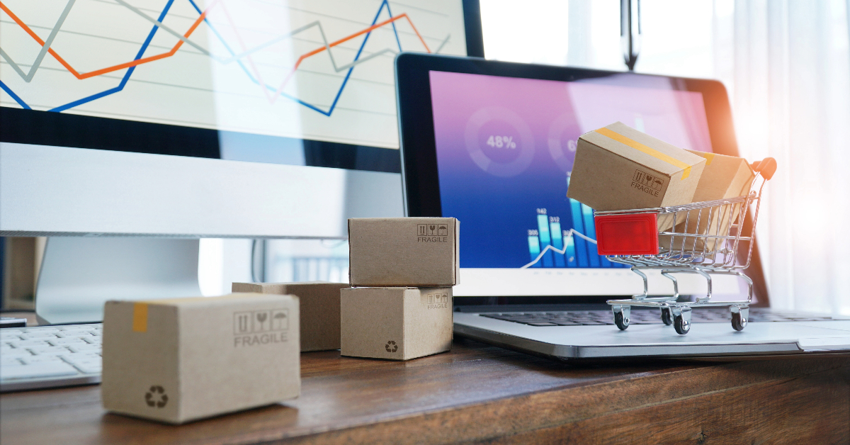 Small cardboard boxes and a miniature shopping cart are placed on a desk in front of computer and laptop screens displaying graphs and charts, showcasing expert tips for optimizing eCommerce operations.