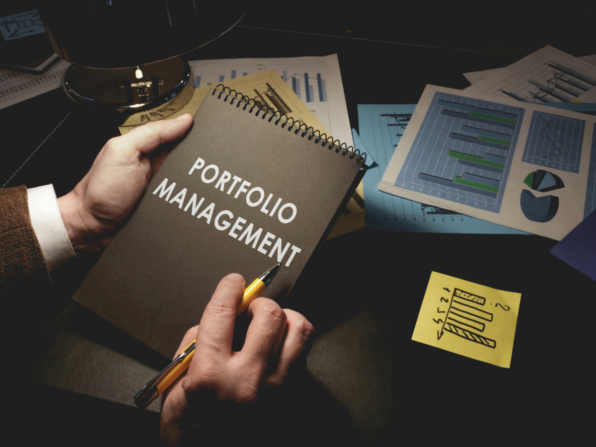 A person holds a notebook labeled "Strategic Portfolio Management" above a desk cluttered with financial charts, graphs, and a sticky note. They hold a pen in their other hand, indicating active engagement in measuring ROI.