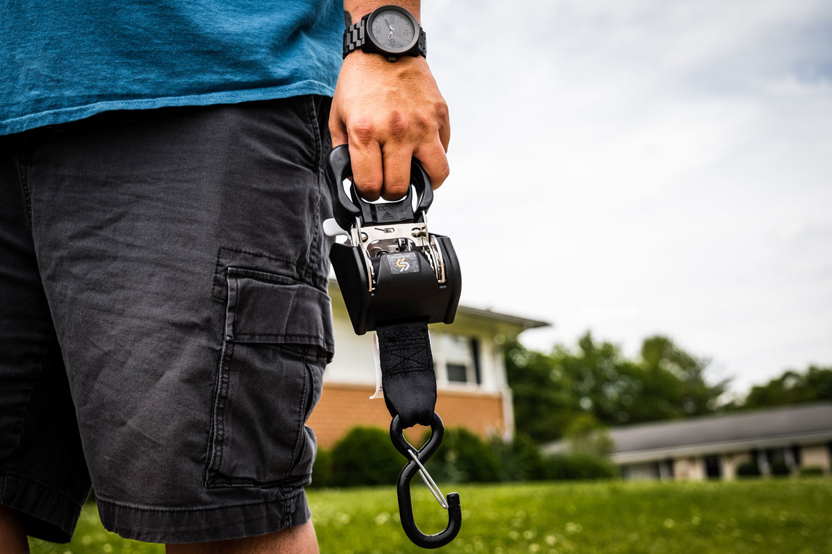 Person wearing a blue shirt and black shorts carrying a black, metal hook and pulley device outdoors near a building, preparing to tie a ratchet strap to secure loads effectively.