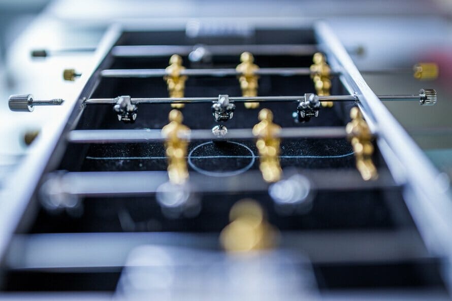Close-up of a foosball table with gold and silver players in formation, reminiscent of a competitive Shopify store setup. The focus is on the central area, showcasing the rods and player figurines against a blurred background.