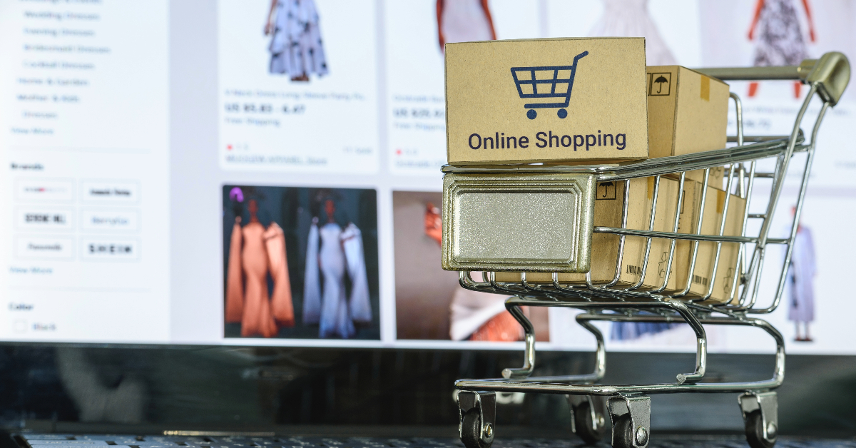 A small shopping cart holding parcels labeled "Online Shopping" is placed in front of a computer screen displaying various clothing items on an online store, illustrating AI for e-commerce and its role in efficient operations.