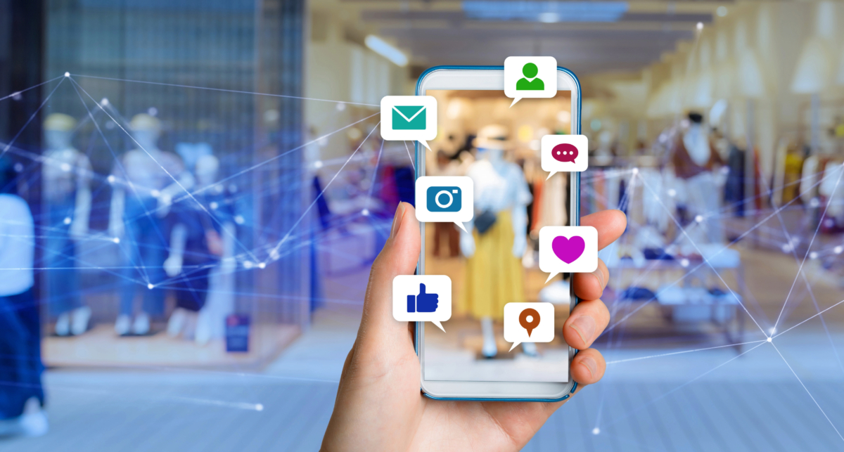 Hand holding a smartphone displaying various social media icons, including Facebook, with a blurred background of a shopping mall showcasing the latest trends.