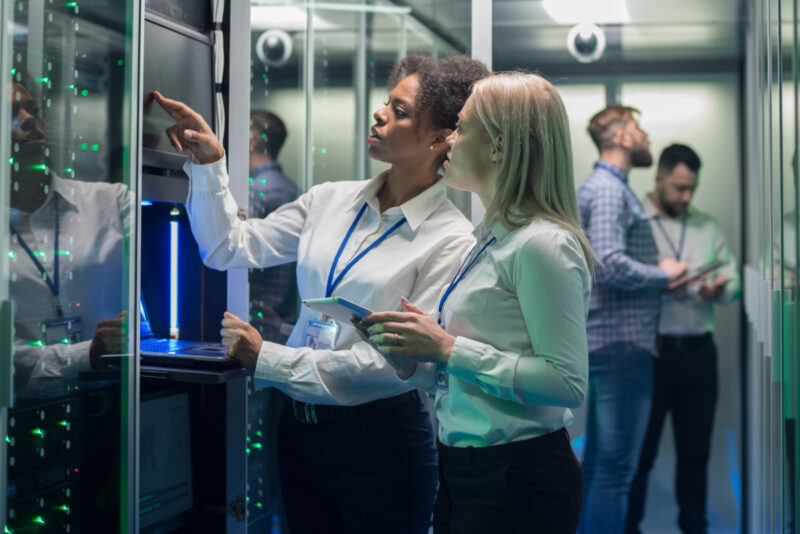 Two women in a server room, one pointing at a screen while the other holds a tablet discussing managed services. Two men are working in the background, ensuring cybersecurity measures to secure your business.