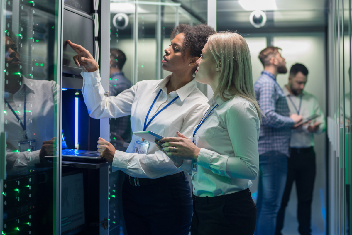 Two women in a server room, one pointing at a screen while the other holds a tablet discussing managed services. Two men are working in the background, ensuring cybersecurity measures to secure your business.