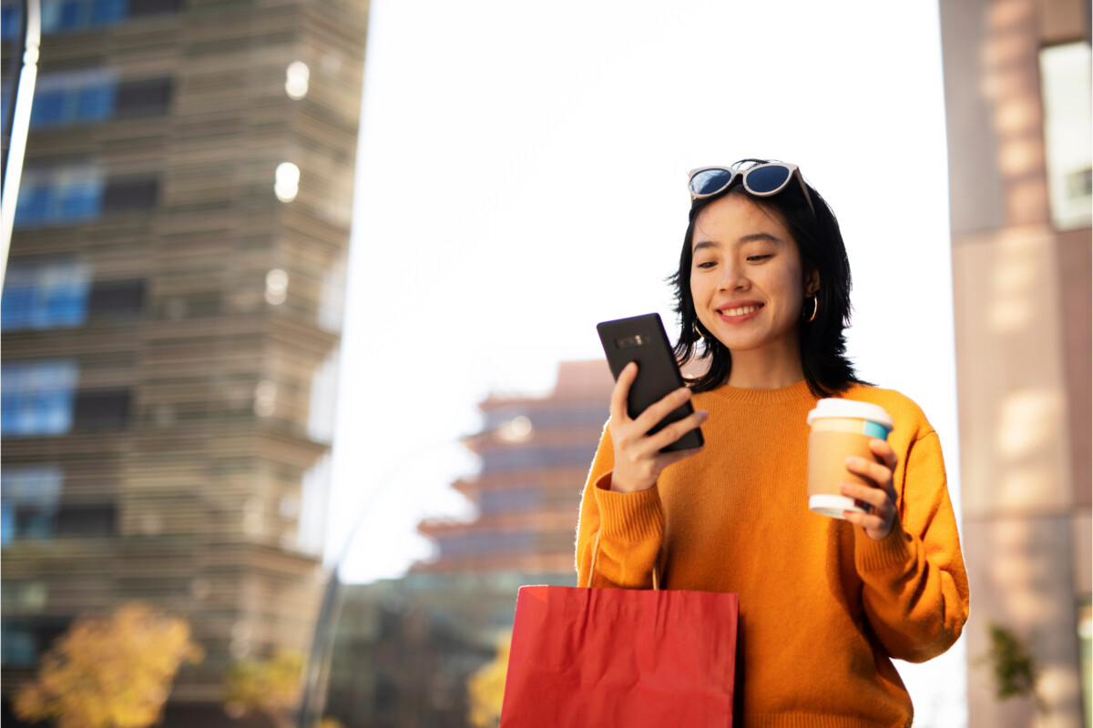 A woman in an orange sweater holds a smartphone and a coffee cup while smiling outdoors in China. She also carries a red shopping bag.