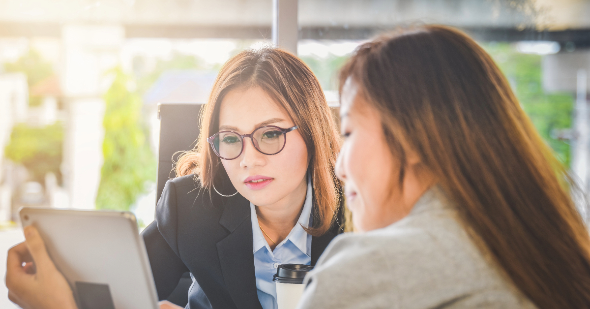 Two women are seated at a table, focused on a tablet. One woman has glasses and is wearing a blazer, exuding the poised confidence required of a CFO, while the other is turned slightly away. They appear to be engaged in an important discussion on how to run a business efficiently.