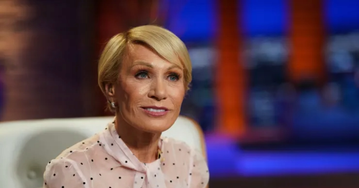 A person with short blonde hair sits in a chair, wearing a light pink blouse with black polka dots. The background is out of focus with hues of blue and orange, reminiscent of an episode where you might meet Barbara Corcoran from Shark Tank.