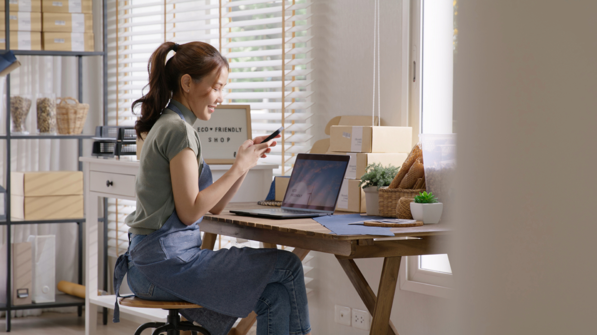 A woman in an apron is sitting at a desk using her phone and laptop in a brightly lit room with boxes and plants around her, perhaps referring to a pricing guide for her Shopify store.