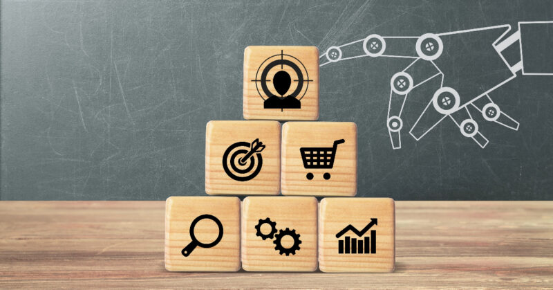 Stacked wooden blocks with various black icons, including a target, shopping cart, gears, bar chart, search icon, and a head in crosshairs, stand in front of a chalkboard drawing of a robotic hand—symbolizing the power of AI-generated content for personalized product recommendations in eCommerce.