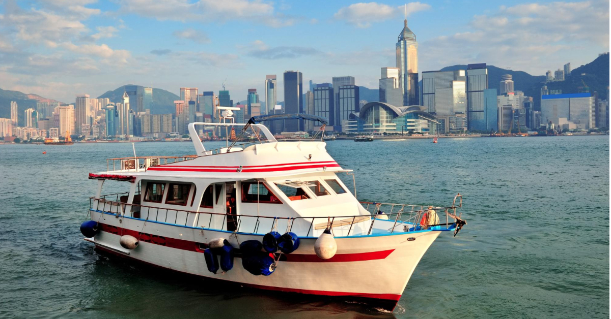 A white boat with red stripes glides on the water, set against a city skyline featuring tall buildings and mountains in the background under a partly cloudy sky. This vessel is the perfect solution for those seeking adventure while offering increasing seating capacity for more passengers.