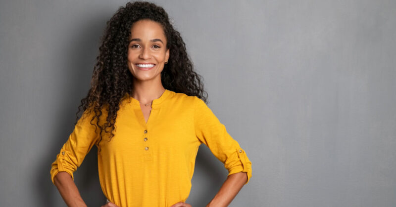 A person with curly hair smiles, wearing a yellow long-sleeve shirt, standing against a plain gray background with hands on hips, looking ready to explore Brazil.