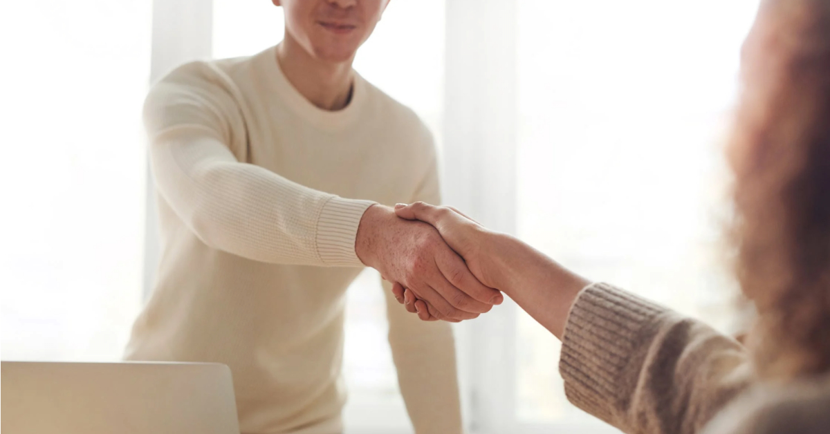 A person in a light sweater is shaking hands with another individual, whose arm is visible in the foreground. The background is softly lit, evoking a sense of professionalism—ideal for those looking for interview tips for an SEO job in 2024.