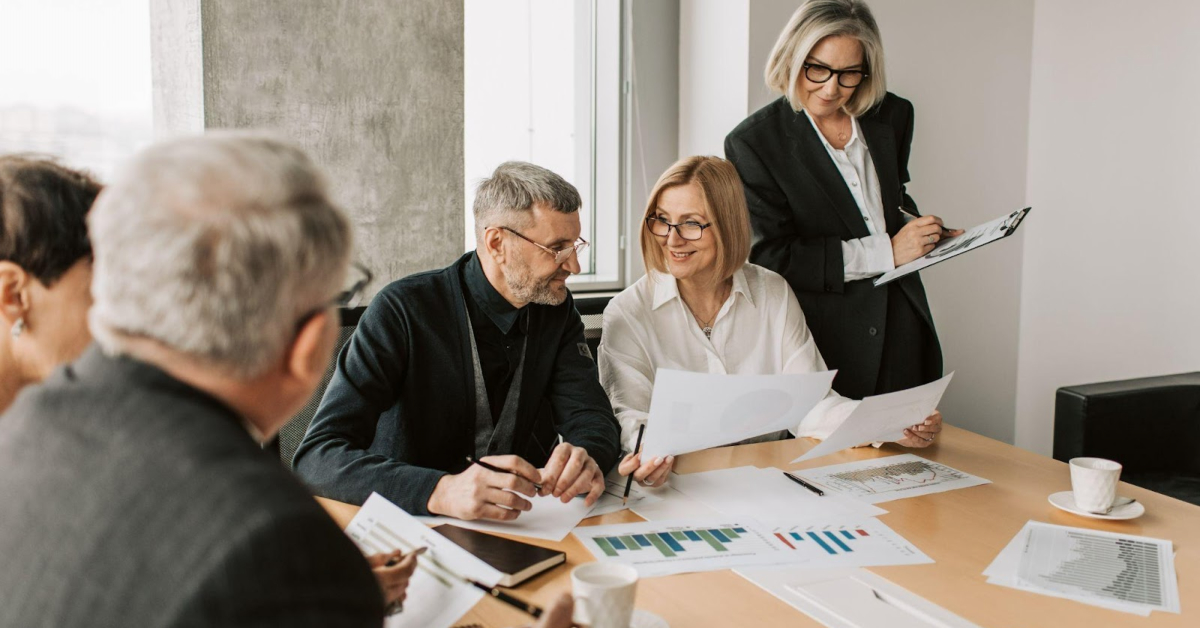 Four people in business attire sit around a table reviewing documents and charts related to sustainable finance, while a fifth person stands holding a clipboard. Coffee cups are placed on the table, reflecting their commitment to ESG principles in every aspect of their meeting.
