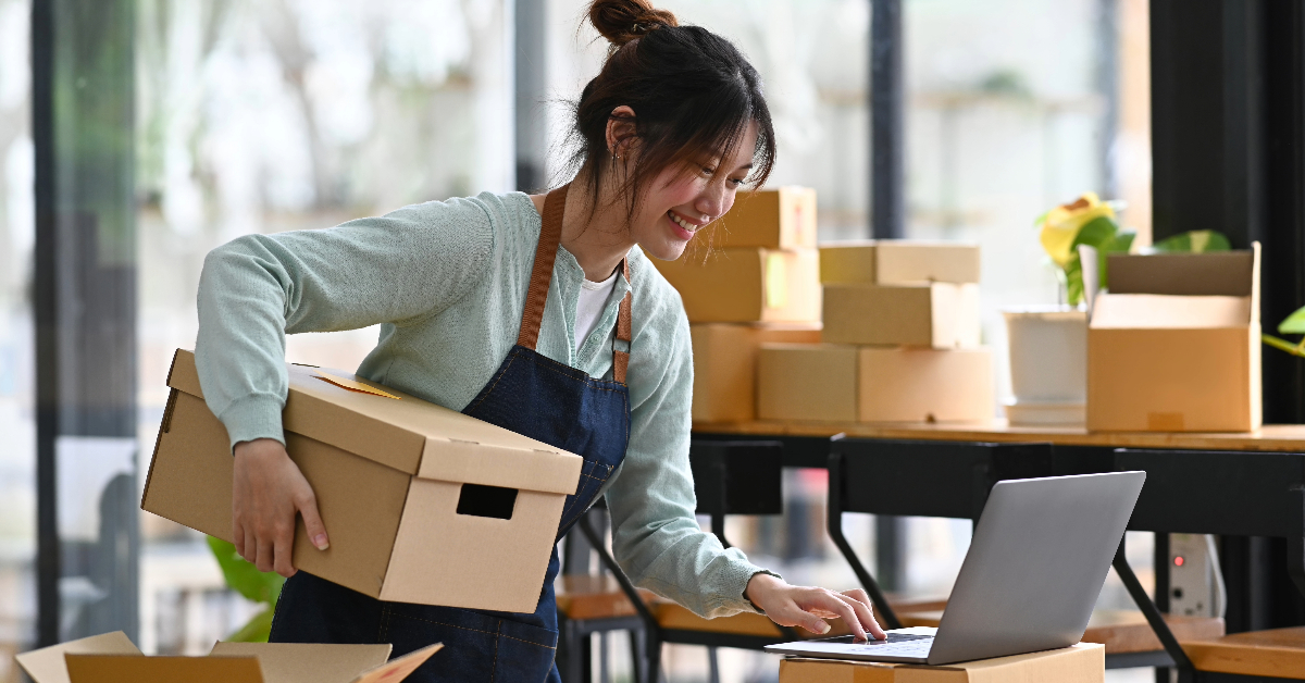 A person in an apron smiles while holding a cardboard box in one hand and working on a laptop with the other, perhaps implementing localization strategies for their business. There are more cardboard boxes in the background.