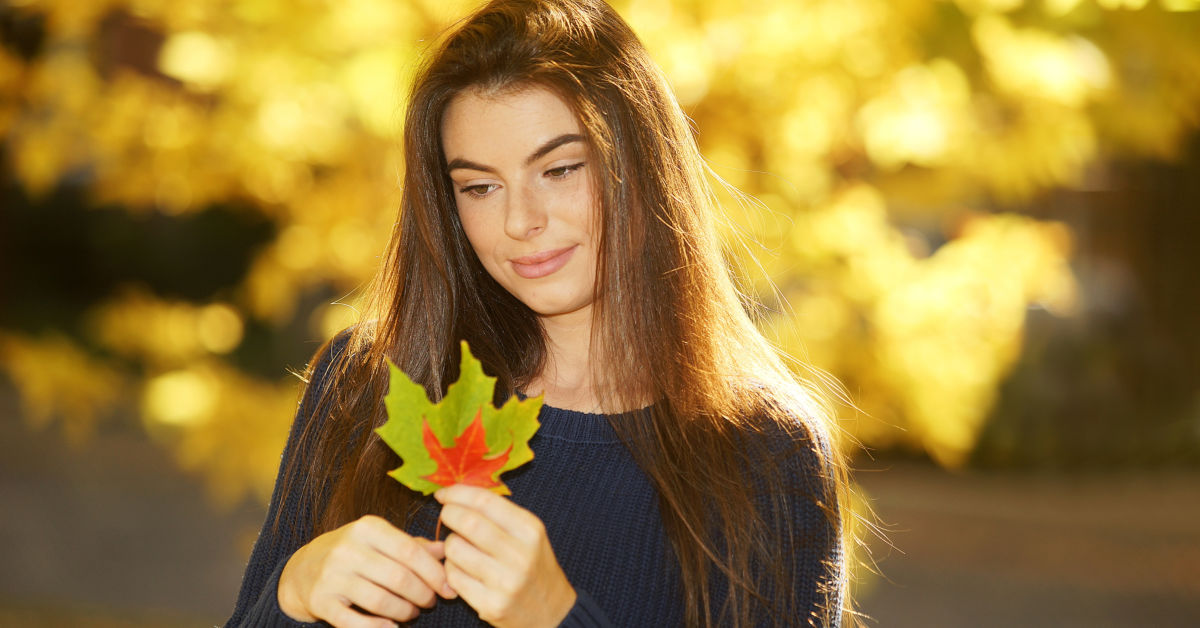 A woman with long brown hair holds a red and green maple leaf, evoking a touch of Canada, with a blurred background of yellow autumn foliage.