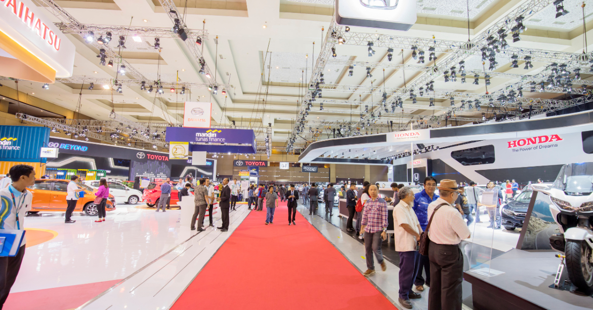 People walking and exploring various custom-built exhibits from brands like Daihatsu, Toyota, and Honda at a car exhibition in a large, well-lit hall with a red carpet and overhead lighting trusses, showcasing strong brand presence and impressive automobile customization.