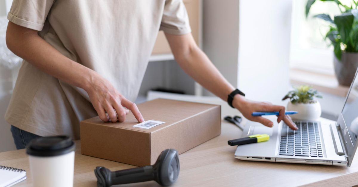 A person scans a barcode on a brown cardboard package while using a laptop on a desk, planning to start an online business in 2023. Nearby are a coffee cup, a barcode scanner, highlighters, and a potted plant.