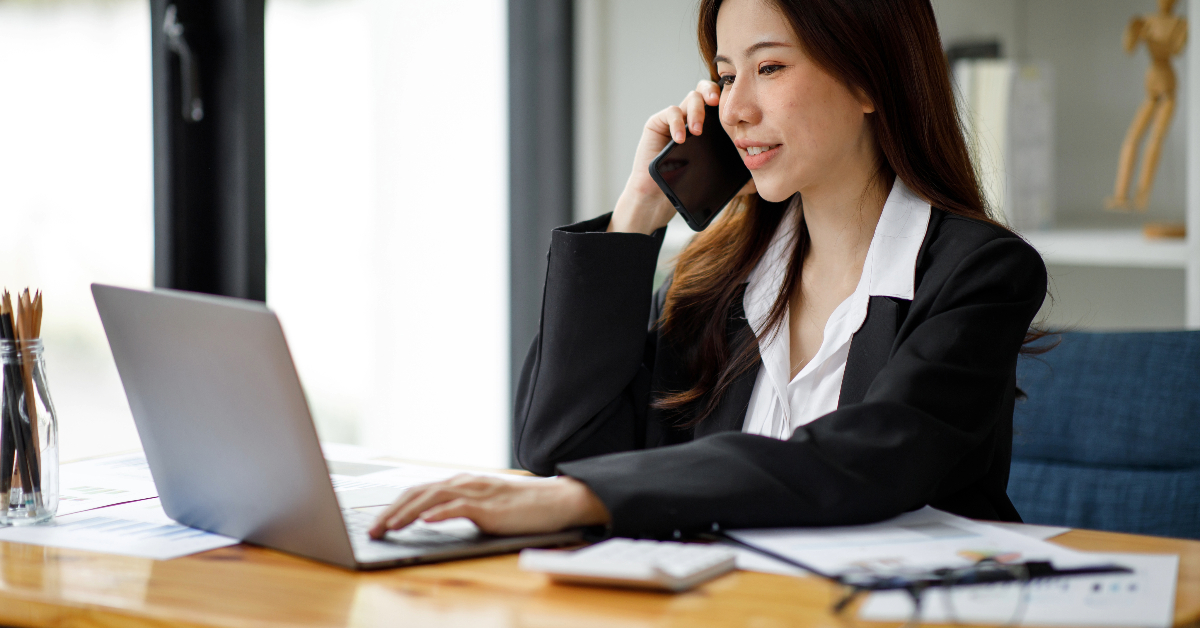 A woman in a black blazer talks on the phone while working on a laptop at a desk, likely discussing strategies for voice-enabled shopping. She appears to be in an office setting.