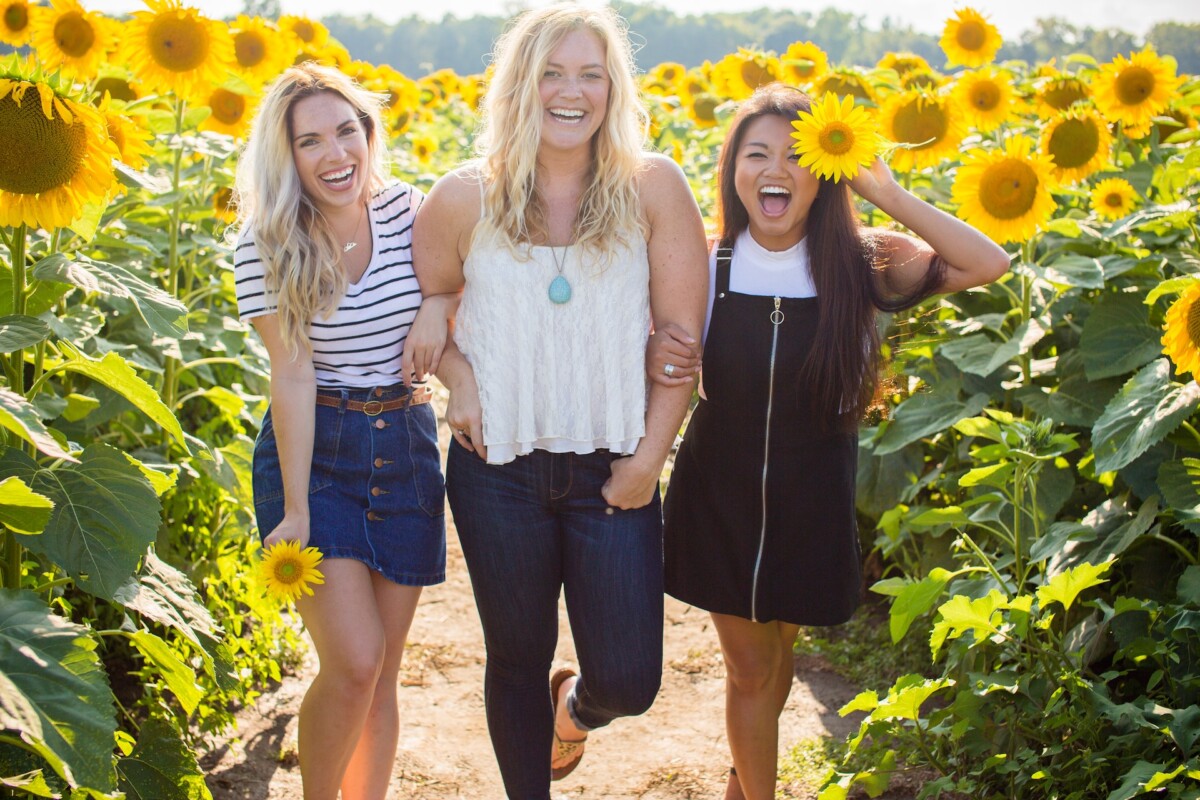 Three women are standing in a field of sunflowers, smiling and appearing to enjoy themselves. The woman in the center, who seems poised and confident, holds a sunflower. Their camaraderie embodies the spirit of breaking barriers as they navigate business challenges together.